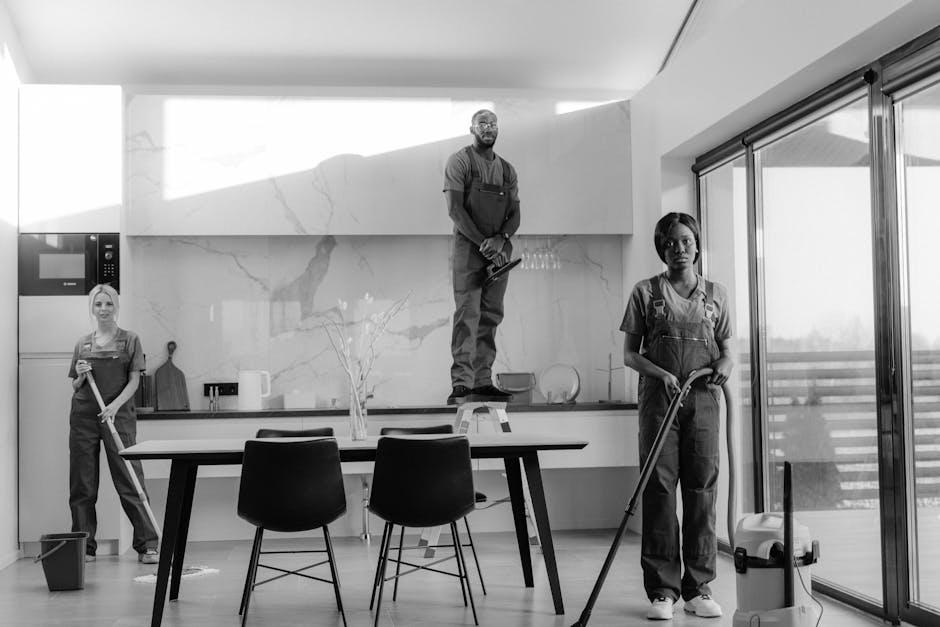 A black-and-white image showing three professional cleaners in a modern, spacious kitchen. One cleaner is standing on a step ladder, wiping or polishing the marble backsplash wall, which has a smooth,