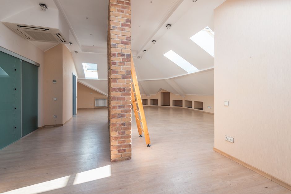 A spacious, bright attic room with a sloped ceiling fitted with skylights, allowing natural light to illuminate the light wooden flooring and beige walls. The room features a brick wall partition with