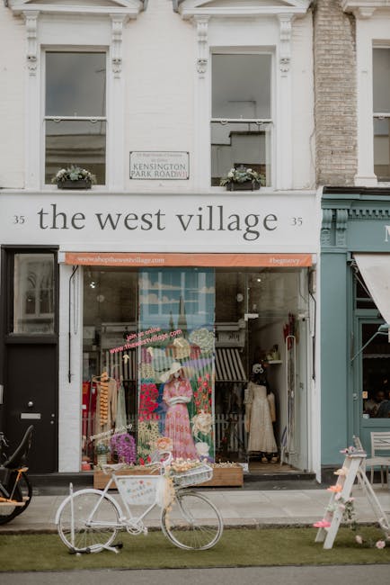 The exterior of a retail shop on Kensington Park Road features a white facade with decorative moldings and two sash windows on the upper floor, each adorned with flower pots. The ground floor showcase