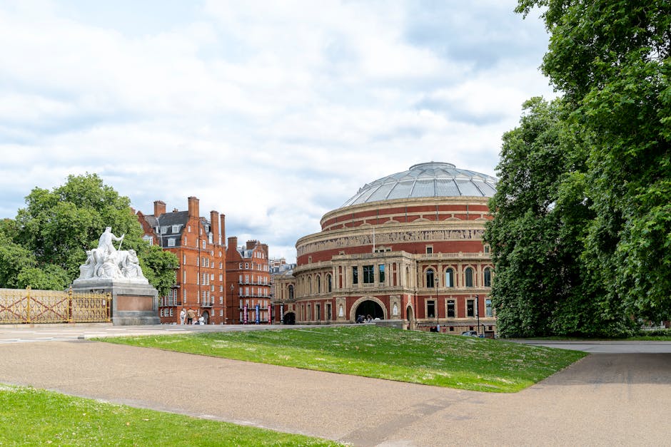 A wide view of the Royal Albert Hall in South Kensington, London, showing the iconic circular red-brick building with a domed roof and arched entrance. The foreground includes a well-maintained grassy