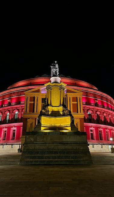 A wide view of the Royal Albert Hall in South Kensington, London, showing the iconic circular red-brick building with a domed roof and arched entrance. The foreground includes a well-maintained grassy
