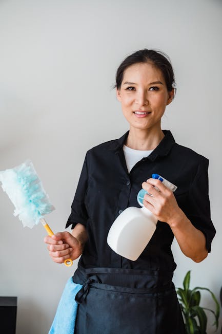 A professional cleaner wearing an orange apron and grey top is standing in a modern living room, holding a vacuum cleaner hose. The vacuum cleaner is yellow and grey, positioned on the carpeted floor.