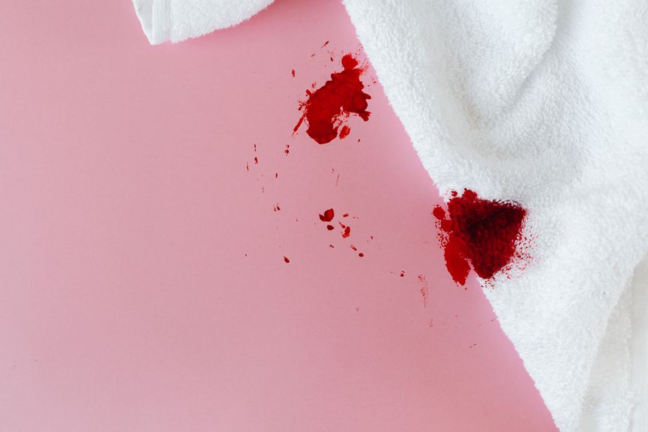 Close-up of a person's hand holding a white cloth with visible red bloodstains, set against a soft pink background. The cloth appears to be a piece of fabric, possibly part of a garment or cleaning ma